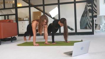 Two young women hold a plank position on a yoga mat with a lap top nearby video