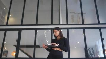 Young woman dressed in black stands while using a laptop in an open space with a large window video