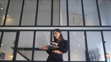 jovem vestida de preto fica enquanto estiver usando um laptop em um espaço aberto com uma grande janela video