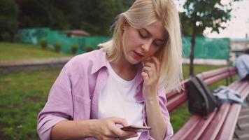 bionda adolescente ragazza si siede su un' panchina mentre utilizzando inteligente Telefono video