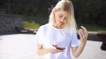 junge blonde frau oder jugendlich blickt bei strahlendem sonnenschein auf telefon-sms video