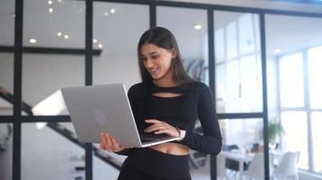 jovem vestida de preto fica enquanto estiver usando um laptop em um espaço aberto com uma grande janela video