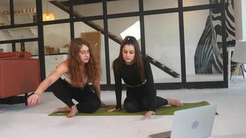 Two young women work out on a yoga mat on the floor with an open laptop nearby video