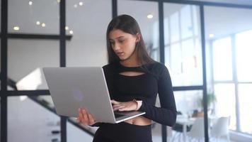 jovem vestida de preto fica enquanto estiver usando um laptop em um espaço aberto com uma grande janela video