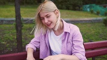 Blonde teenage girl smiles at phone and then to camera while seated at a park bench as a breeze blows