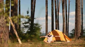 Young woman sits on a yoga mat outside a yellow tent at camp site