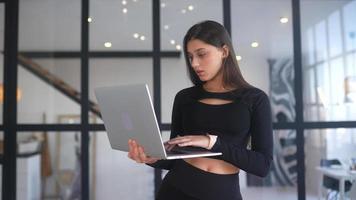 giovane donna vestito nel nero sta mentre utilizzando un' il computer portatile nel un Aperto spazio con un' grande finestra video