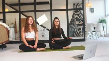 Two young women work out on a yoga mat on the floor with an open laptop nearby video