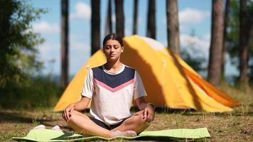 Young woman sits on a yoga mat outside a yellow tent at camp site video