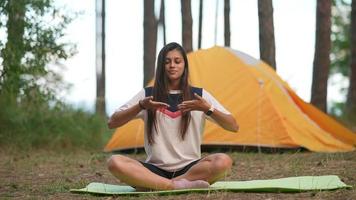 Young woman sits on a yoga mat outside a yellow tent at camp site