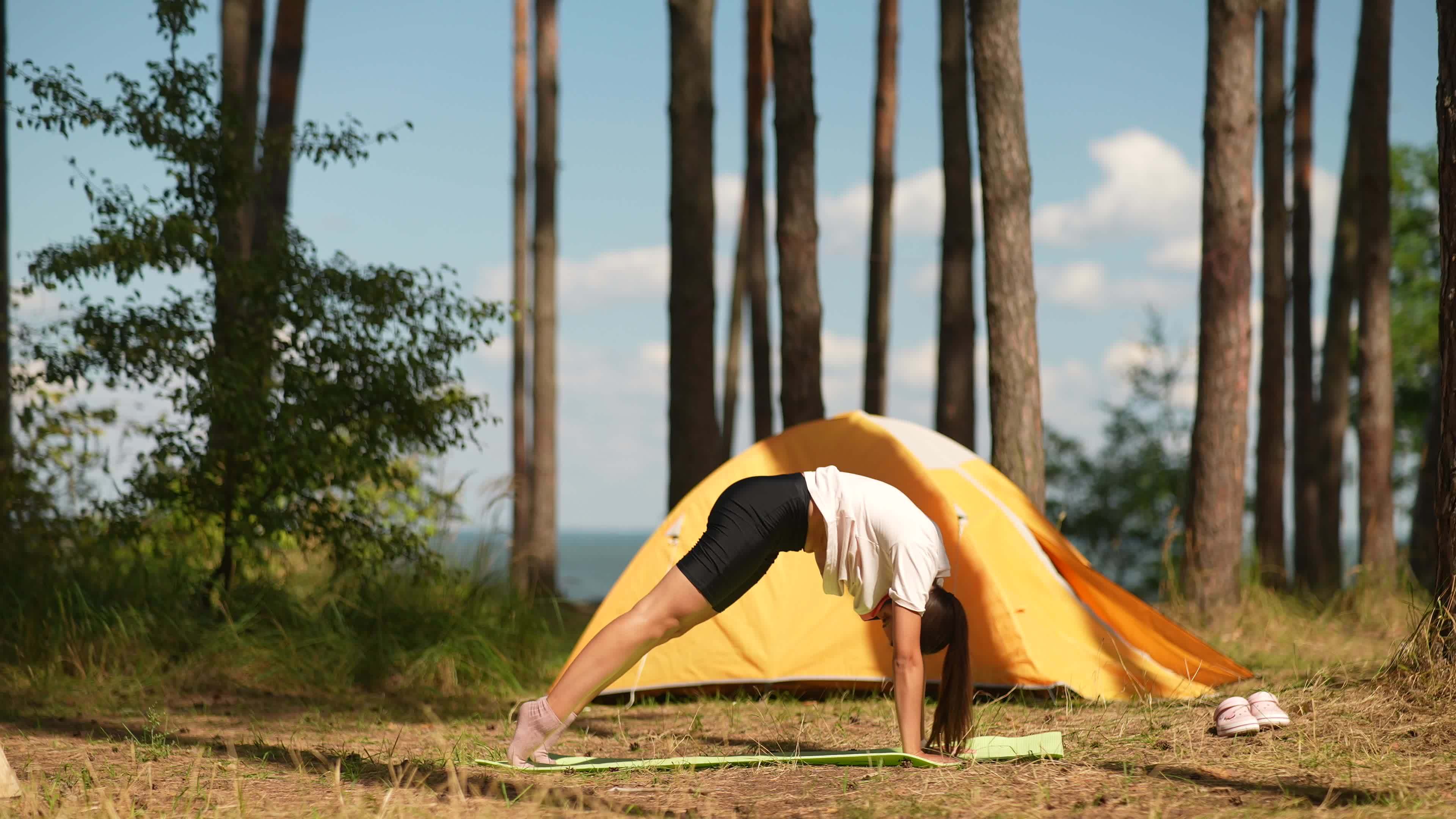 Young woman stretches on a yoga mat outside a yellow tent at camp site