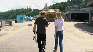 Two teenagers walk toward carousel carrying skateboard video