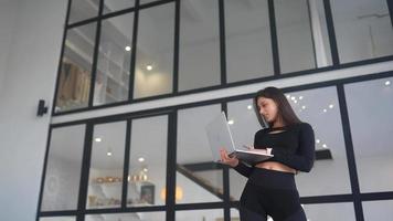 Young woman dressed in black stands while using a laptop in an open space with a large window video