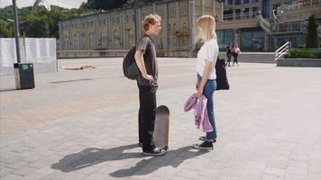 Teen boy and girl with backpacks and skateboard stand and talk in sunny outdoor city space video
