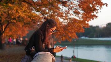 donna sembra a inteligente Telefono mentre nel un' parco nel autunno video