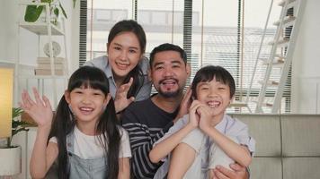 Happy Asian Thai parents, and children looking and talking to a camera, online video call with family via internet on sofa in home living room, lovely weekend, and domestic wellbeing lifestyle.
