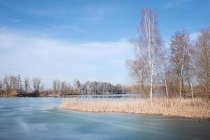 In winter, bare birches and other trees and bushes stand against a blue sky by a partially frozen lake in Bavaria photo