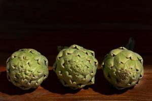 Three artichokes lie next to each other on a wooden base in front of a dark background with space for text photo