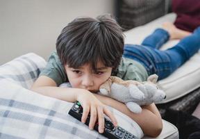 Dramatic portrait Lonely kid sad face holding remote control sitting on couch,child sitting on sofa in living room with looking out deep in thought, Boy bored stay at home alone. photo