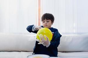 niño lindo con guante de plástico protector desechable y sosteniendo un globo amarillo, niño preparando un proyecto científico sobre carga estática, niño mirando curiosamente el experimento científico, concepto de educación en el hogar foto