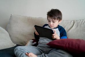 Indoor portrait  kid sitting on sofa watching cartoon on tablet, Cute boy playing game on touch pad, Child having fun and relaxing on his own in living room, New normal lifestyle photo