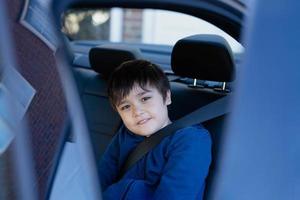 Cinematic portrait boy siting in safety car seat looking at camera with smiling face,Child sitting in the back passenger seat with a safety belt, School kid traveling to school by car.Back to school photo