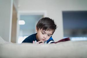 niño de tiro sincero sentado en el sofá viendo dibujos animados en la tableta, niño lindo jugando en el panel táctil, niño divirtiéndose y relajándose solo en la sala de estar, nuevo estilo de vida normal foto