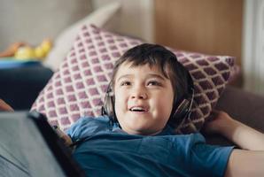 Happy kid wearing headphone looking up watching TV, Smile boy lying on sofa listening  music on Tablet, Child with smiling face having fun playing games at home. New normal life style photo