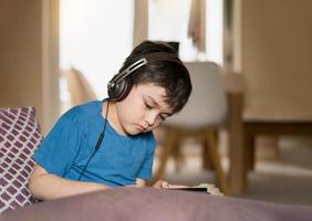 Authentic portrait Kid sitting on sofa watching cartoons on tablet,Yong boy playing game on touch pad, Child lying on couch wearing headphoes listening to music or relaxing on his own in living room photo