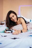 chica divertida acostada en la cama y jugando videojuegos, sosteniendo el controlador foto