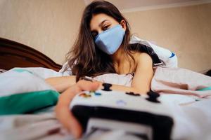 Girl in protective mask lying in bed and holding controller photo