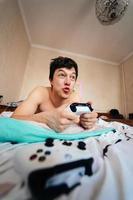 Guy lying in bed and playing video game, holding controller photo