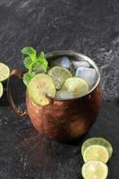 Cóctel alcohólico de mula de Moscú en taza de cobre con hielo, menta y limón en rodajas sobre fondo de piedra negra. foto