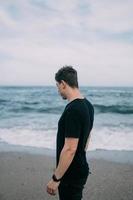 Smiling guy in a black T-shirt stands on the sandy seashore. photo