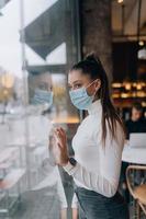 mujer joven con mascarilla parada frente a las ventanas en el café. foto