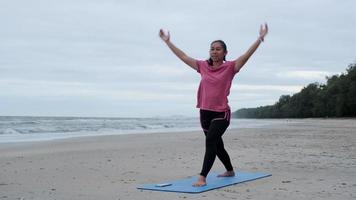 mujer asiática practicando yoga en la orilla del mar. joven hermosa mujer estirándose y haciendo yoga al aire libre en una alfombra de yoga azul en la playa. relajante en la naturaleza video