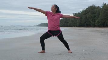 mulher asiática praticando ioga à beira-mar. bela jovem esticando e fazendo ioga ao ar livre no tapete de ioga azul na praia. relaxando na natureza