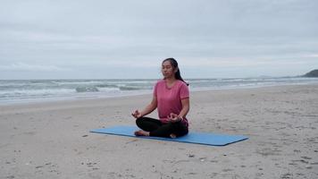 mujer asiática practicando yoga en la orilla del mar. hermosa mujer meditando alrededor de la playa del mar al amanecer para la salud. relajante en la naturaleza video