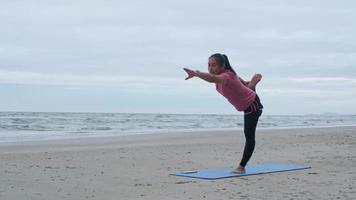 Asian woman practicing yoga at seashore. Young beautiful woman stretching and doing yoga outdoors on blue yoga mat on beach. relaxing in nature video