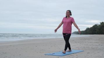 mujer asiática practicando yoga en la orilla del mar. joven hermosa mujer estirándose y haciendo yoga al aire libre en una alfombra de yoga azul en la playa. relajante en la naturaleza video