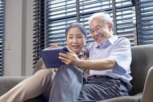 pareja asiática mayor en edad de jubilación mirando una foto nostálgica usando una tableta digital mientras se sienta en el sofá de su casa de retiro para el concepto de actividades de ancianos y pensiones
