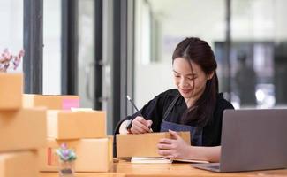 Young beautiful happy asian business woman owner of SME online using laptop receive order from customer with parcel box packaging at her startup home office, online business seller and delivery photo