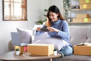 Asian woman photographing parcel boxes with smartphone to deliver goods to customers online on the Internet, starting a small business owner's e-commerce concept. photo