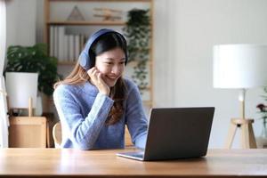 Cheerful beautiful Asian woman wearing headphones and look to digital tablet at video calling meeting and study online on the Internet.