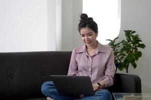 Mujer asiática independiente de negocios joven que trabaja en la computadora portátil que controla las redes sociales mientras está acostado en el sofá cuando se relaja en la sala de estar en casa. estilo de vida de las mujeres de etnia latina e hispana en el concepto de casa. foto