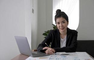 Beautiful smiling Asian businesswoman hand holding pen working using tablet at office. Looking at camera. photo