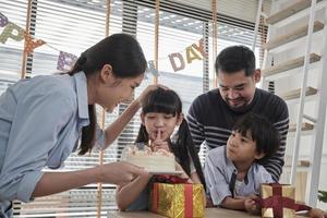 feliz familia tailandesa asiática, hija pequeña sorprendida, pastel de cumpleaños y regalo encantador, niña sopla velas, reza y celebra alegremente la fiesta con los padres juntos, estilo de vida especial de eventos domésticos. foto