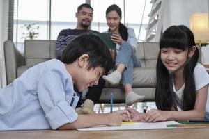 Asian Thai siblings are lying on living room floor, drawing homework with colored pencils together, parents leisurely relax on a sofa, lovely happy weekend activity, and domestic wellbeing lifestyle. photo