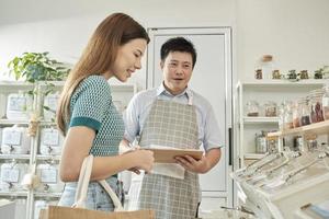 Asian male shopkeeper describes natural organic products to woman customer in refill store, zero-waste and plastic-free grocery, eco environment-friendly, sustainable lifestyles with a reusable shop. photo