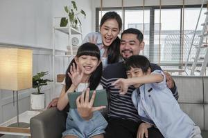 Happy Asian Thai parents, and children online video call with family via internet on sofa in home living room, lovely weekend, and domestic wellbeing lifestyle with internet technology mobile phone. photo